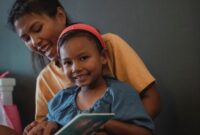 Seorang ibu sedang membacakan buku cerita bergambar kepada anaknya di ruang keluarga yang hangat sebelum tidur. Aktivitas mendongeng ini menjadi sarana efektif untuk mempererat ikatan batin sekaligus menanamkan nilai moral kearifan lokal sejak dini.