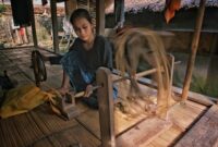 Seorang perempuan perajin di Sumba Timur memintal benang menggunakan alat tenun kayu tradisional di pekarangan rumah adatnya.